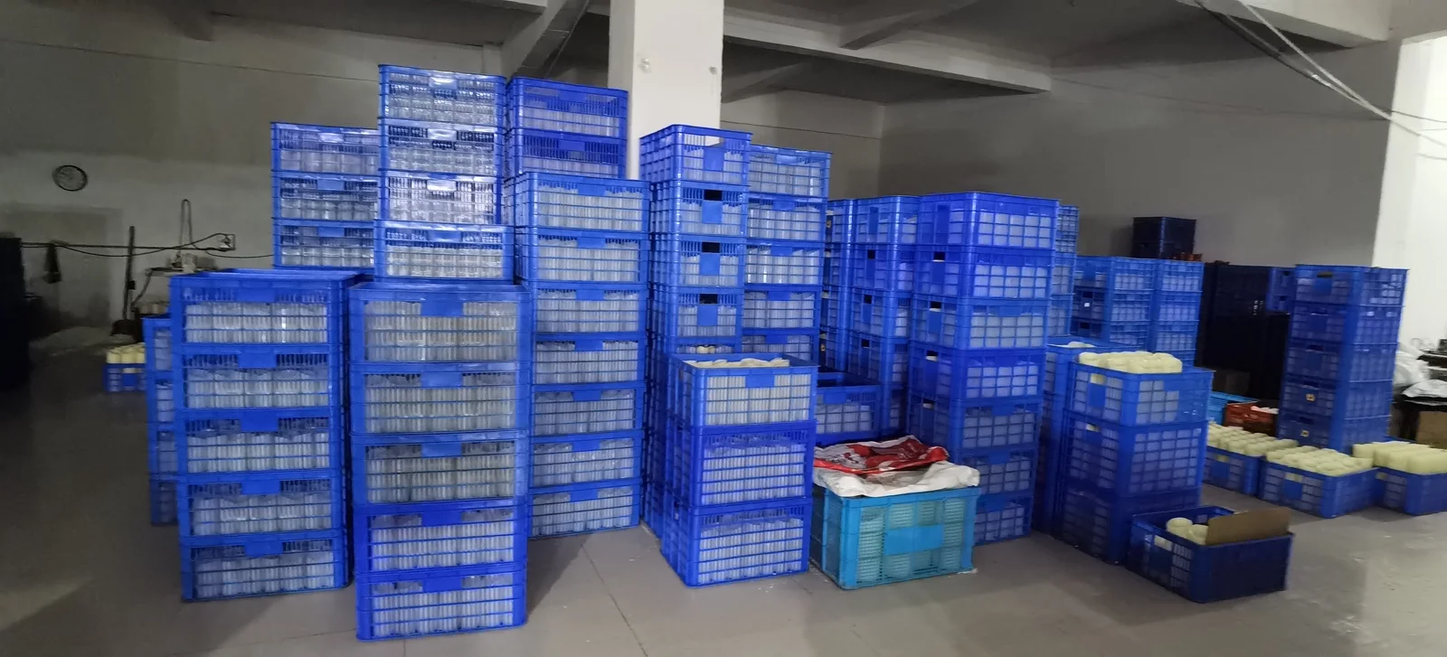 Blue stacking crates filled with candles in clear cups piled high across a factory warehouse floor, with trays of unpoured wax at the right