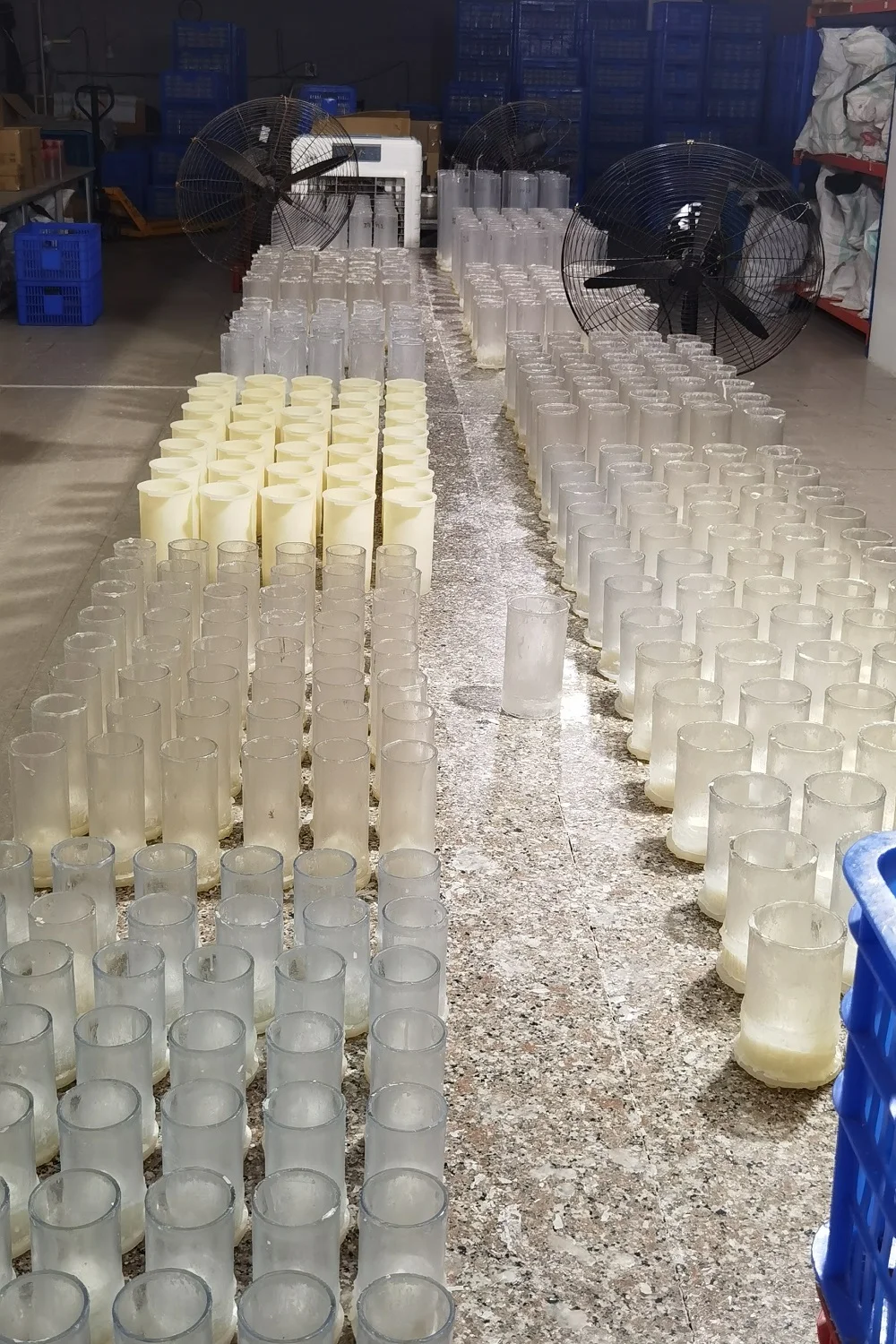 Rows of white candle bodies and clear glass candle components arranged on a factory worktable with blue crates in the background