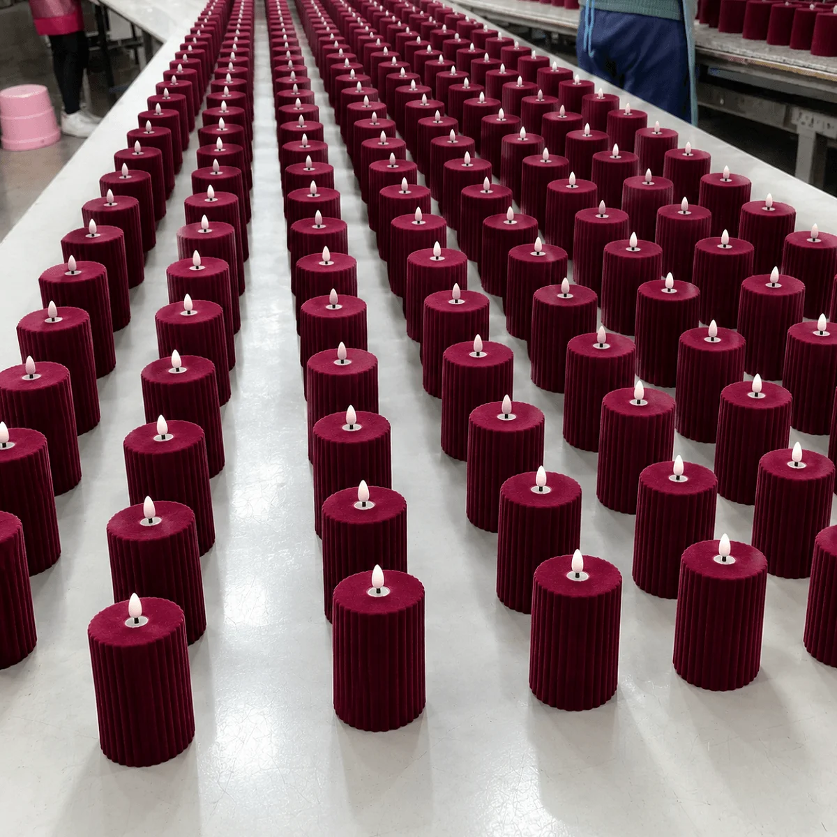 Hundreds of wine-red fluted flameless pillar candles lined up in rows across a white factory finishing table