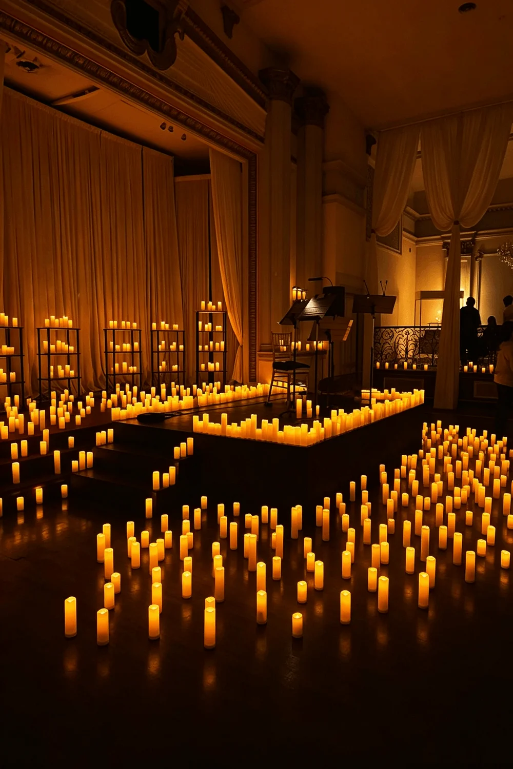 Hundreds of warm glowing flameless pillar candles arranged across the floor and stage of a concert hall