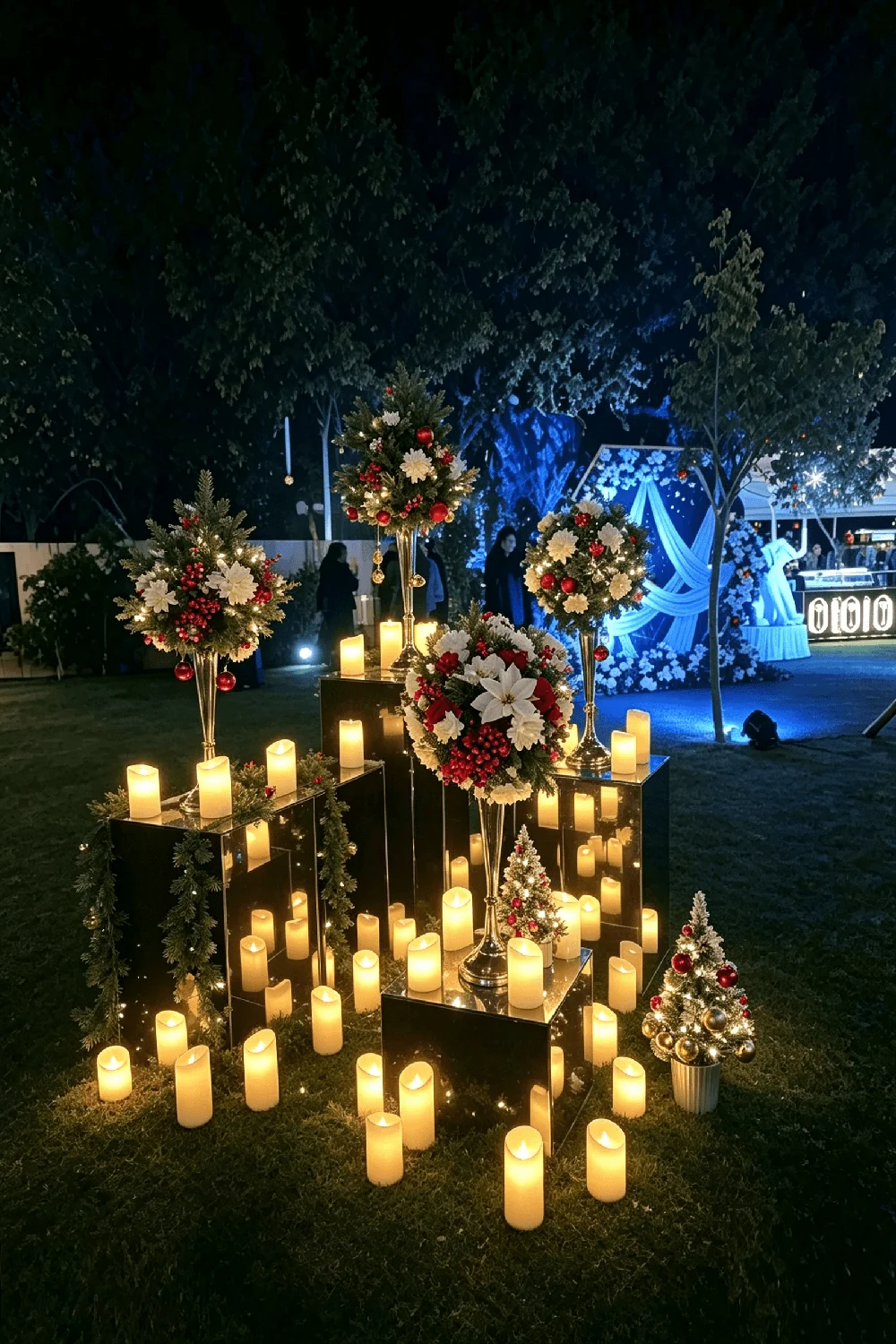 Dozens of ivory flameless pillar candles massed over mirrored plinths and grass on a night lawn, beneath tall stands of white flowers and red berries