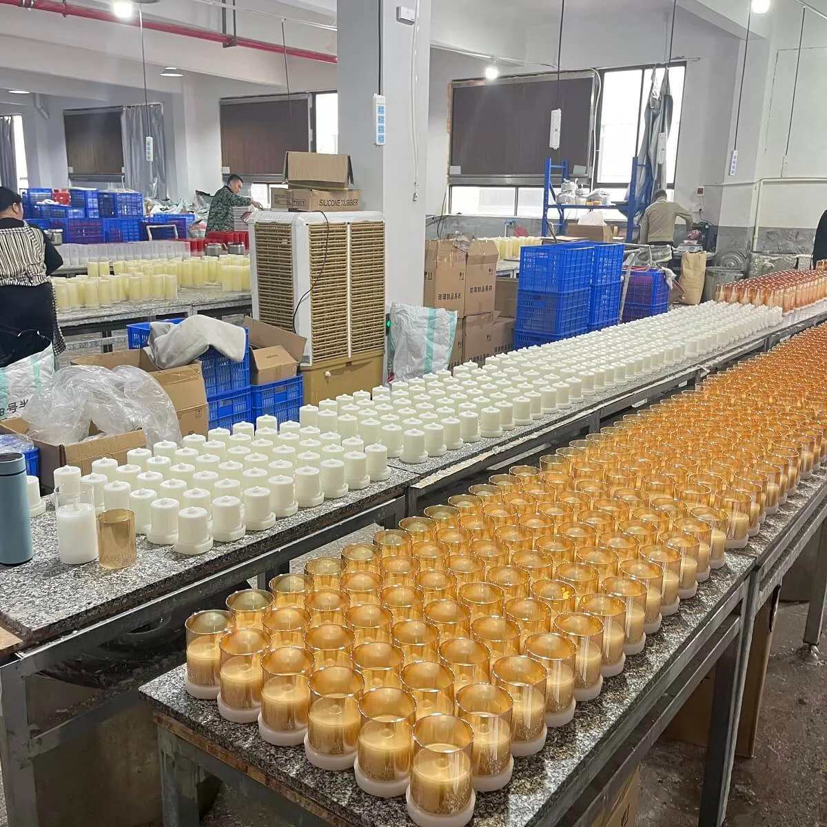 Long factory worktables lined with rows of white LED candle bodies and amber glass votive shells, with staff assembling more in the background