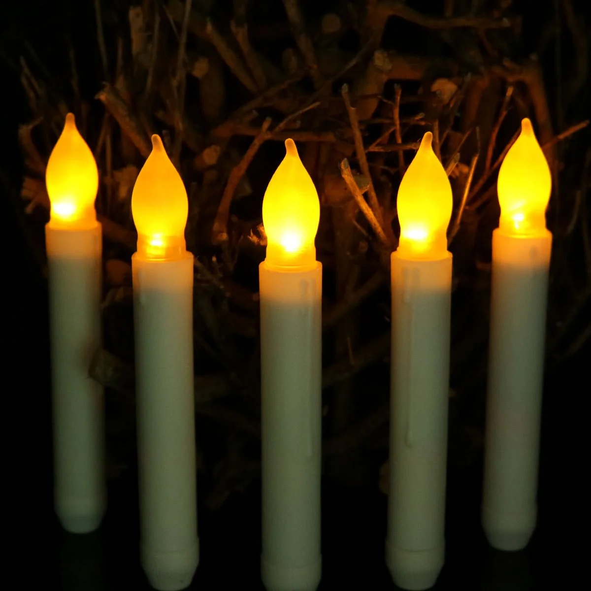 Five ivory flameless taper candles with amber flickering flame tips standing against a background of bare twigs