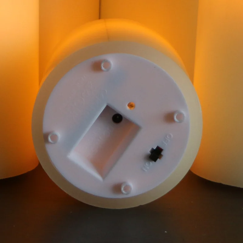 Close-up of the underside of a flameless candle showing its recessed charging contact, a small amber indicator light and an on-off switch, backlit by two glowing candles