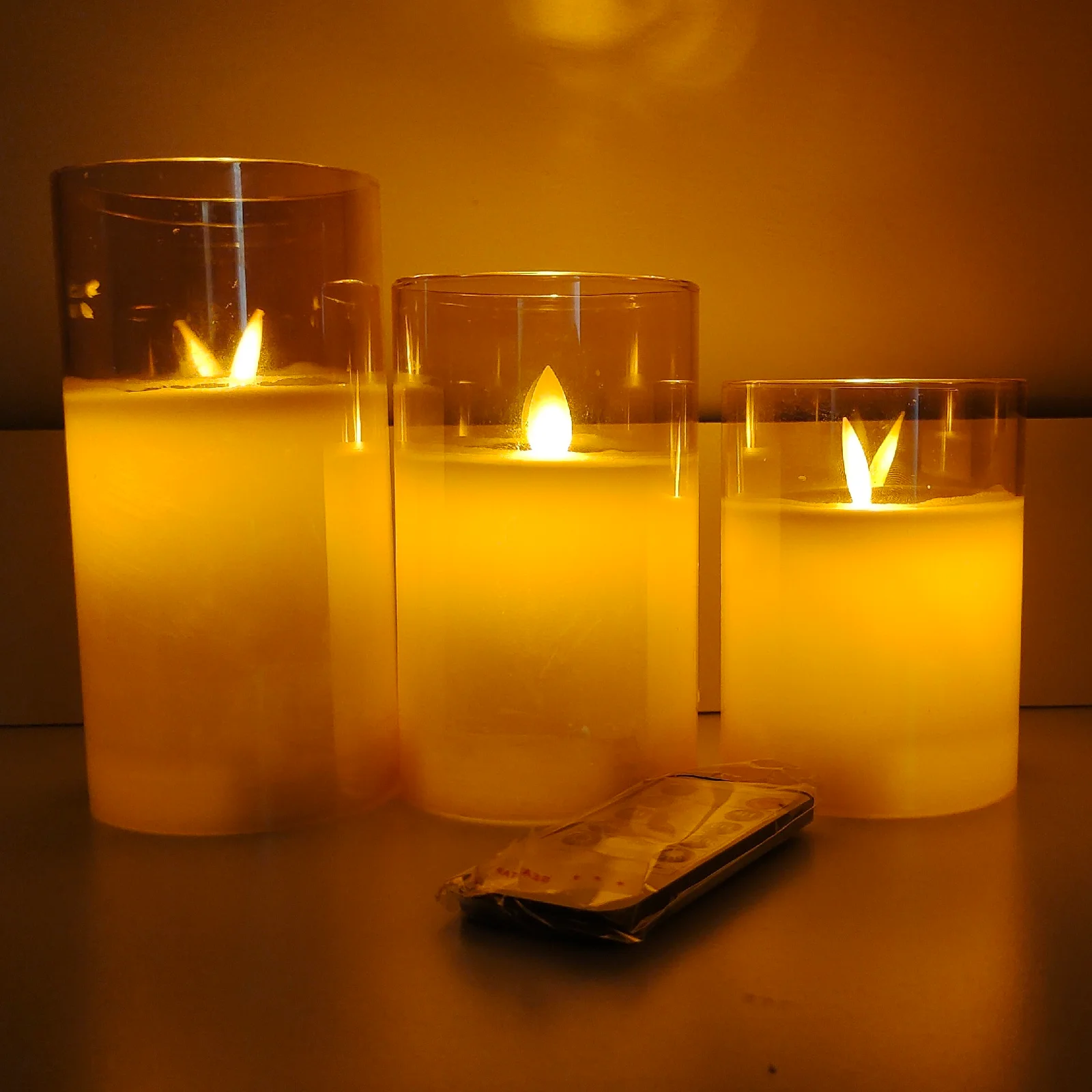 Three sizes of ivory flameless pillar candles lit inside clear glass cylinders, with a remote handset lying in front of them